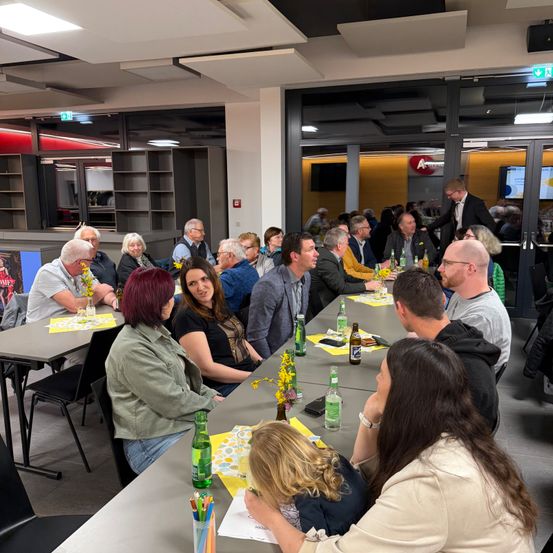 Eine Gruppe von Menschen sitzt an einem Tisch, einige mit Getränken und gelben Blumen. Die Umgebung scheint ein modernes Büro oder Konferenzraum mit Glaswänden zu sein.