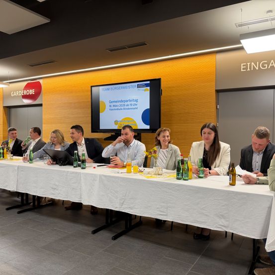 Eine Gruppe von Menschen sitzt an einem Tisch mit einem Monitor, der den Text 'Gemeindeparteitag' anzeigt. Auf dem Tisch befinden sich Getränke und Laptops. Sie scheinen etwas zu besprechen.