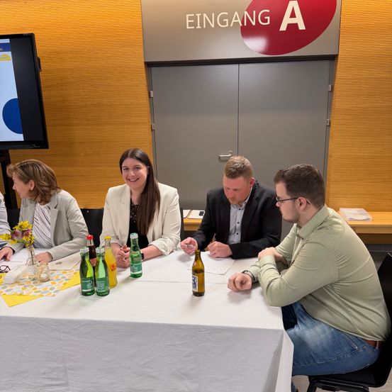 Eine Gruppe von Menschen sitzt um einen Tisch mit Getränken. Das Schild oben lautet 'Eingang A'. Sie scheinen eine Besprechung zu haben.