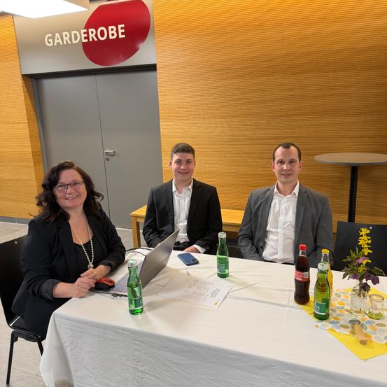Drei Personen sitzen an einem Tisch mit Laptop und Getränken. Eine Frau und zwei Männer. Hinter ihnen eine Tür mit der Aufschrift 'Garderobe'.