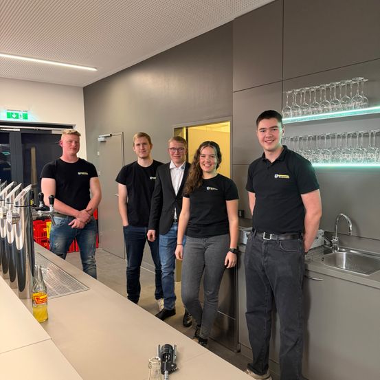 Fünf Menschen in schwarzen T-Shirts stehen in einer Küche. Sie scheinen für ein Foto zu posieren. Dahinter ist ein Waschbecken mit einem Wasserhahn. Auf dem Tresen steht eine Flasche. An der Wand befinden sich Regale mit Weingläsern.