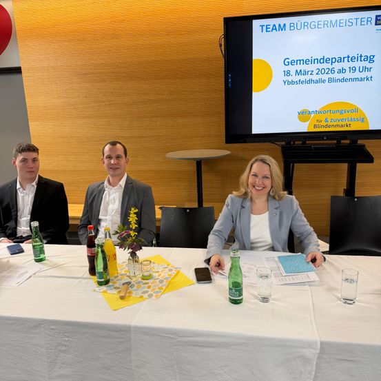 Drei Personen sitzen an einem Tisch mit verschiedenen Flaschen und Papieren. Hinter ihnen befindet sich ein Bildschirm mit einer Präsentation über eine Gemeinschaftsveranstaltung am 18. März 2026.