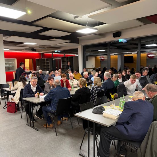 Eine Gruppe von Menschen sitzt an Tischen in einem modernen, gut beleuchteten Raum. Flaschen, Tassen und Papiere liegen auf den Tischen. Der Raum hat eine rote Wand, große Fenster und Deckenleuchten.