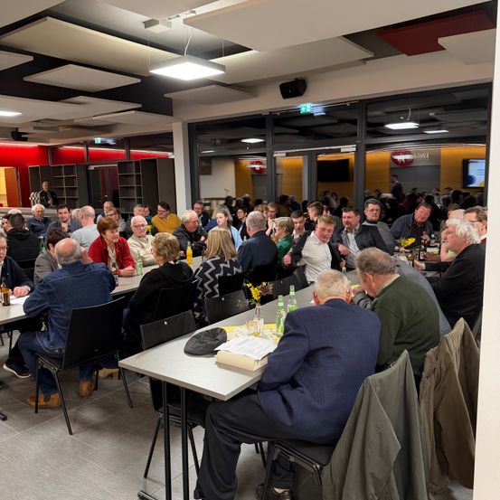 Eine große Gruppe von Menschen, hauptsächlich Männer, sitzen um Tische herum in einem Raum. Einige unterhalten sich, während andere sich auf ihre Getränke und Speisen konzentrieren. Der Raum ist gut beleuchtet und es gibt eine Glaswand mit einem Ausgangsschild.