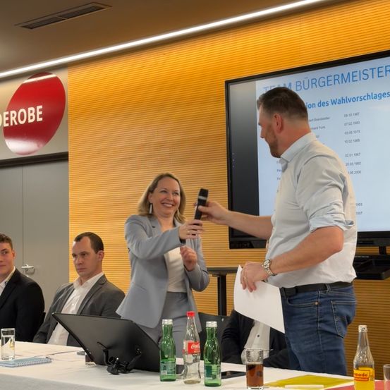 Ein Mann mit einem Telefon in der Hand steht neben einer Frau, beide lächeln. Sie befinden sich in einem Konferenzraum mit einer Präsentationsleinwand dahinter. Andere Personen sitzen am Tisch, und ein Laptop, Getränke und Papiere sind auf dem Tisch.