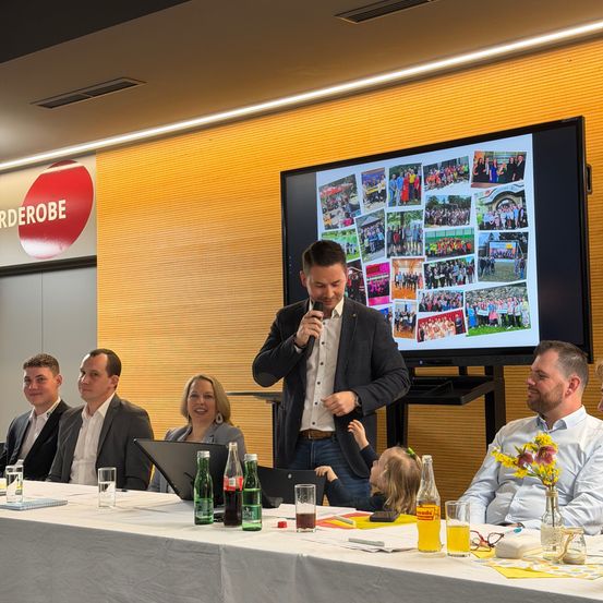 Ein Mann spricht in ein Mikrofon an einem Rednerpult in einem Konferenzraum. Ein Bildschirm mit Fotos befindet sich hinter ihm. Menschen sitzen an einem Tisch und lächeln ihn an.