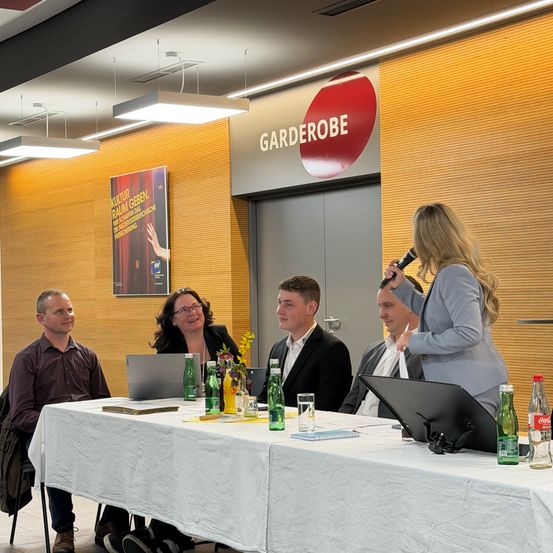 Fünf Personen sitzen an einem Tisch in einem Konferenzraum, eine Frau spricht in ein Mikrofon. Der Raum hat ein Banner mit dem Wort Garderobe.