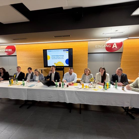 Eine Gruppe von Menschen sitzt um einen Tisch herum mit Flaschen, einem Laptop und einer Projektionsleinwand, die eine Präsentation über eine Gemeindeveranstaltung am 13. März 2026 zeigt.