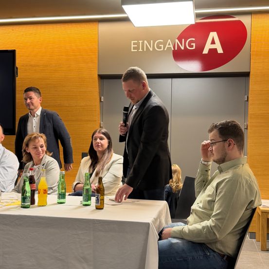 Ein Mann mit Mikrofon spricht bei einer Konferenz. Mehrere Personen sitzen vor ihm an einem Tisch. Flaschen stehen auf dem Tisch. Das Schild lautet 'Eingang A'.