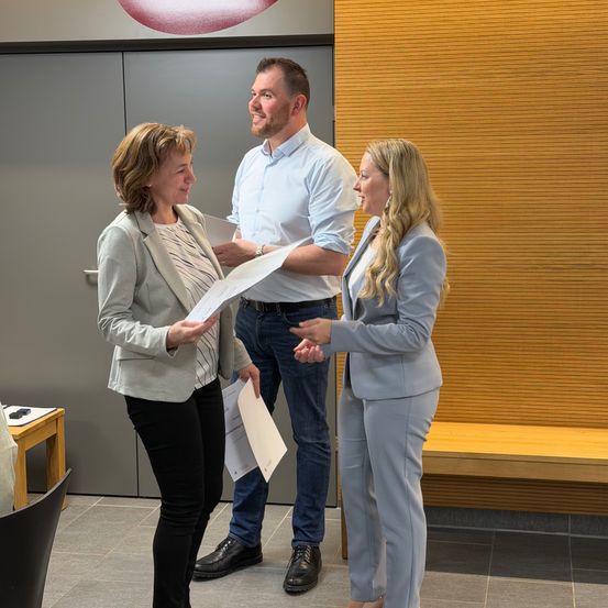 Drei Personen stehen in einem modernen Büro. Zwei Frauen unterhalten sich, während der Mann Papiere hält und lächelt. Der Raum ist mit gefliesten Böden, Holzbänken und einer grauen Tür ausgestattet.