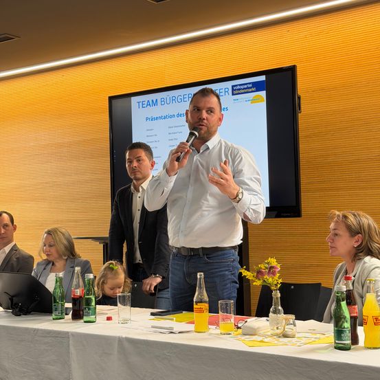 Ein Mann spricht in ein Mikrofon bei einer Präsentation vor einem Bildschirm. Dahinter sitzen eine Frau und ein Kind. Zwei Männer stehen an der Seite, und andere sitzen an einem Tisch mit Getränken.