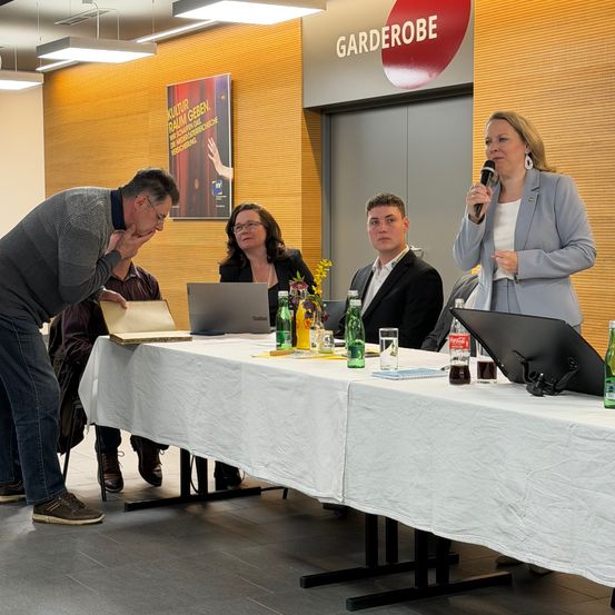 Eine Frau hält ein Mikrofon vor einem Tisch mit Laptops, Getränken und Blumen. Ein Mann beugt sich über den Tisch und schaut sich etwas an. Andere Leute sitzen um den Tisch herum. Ein Poster und Lampen sind an der Wand.