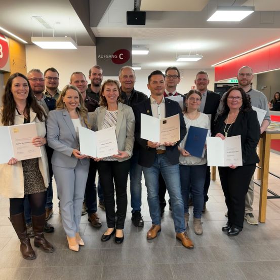 Eine Gruppe von Fachleuten posiert für ein Foto im Büro, während sie Urkunden halten und lächeln. Sie sind in Business-Kleidung gekleidet und scheinen eine Leistung zu feiern.