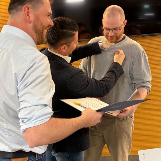 Drei Männer stehen drinnen, einer hält ein Zertifikat, der andere legt eine Medaille auf die Brust des dritten Mannes. Der Mann mit der Medaille lächelt, während die anderen zusehen.