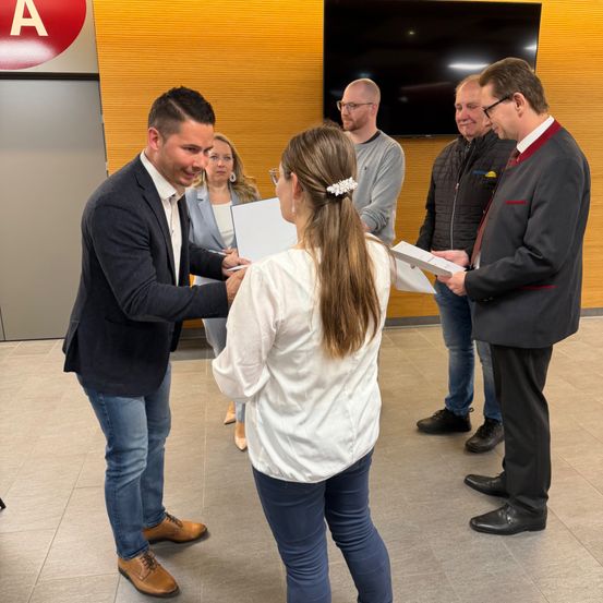Ein Mann in einem Anzug übergibt einer Frau mit Pferdeschwanz ein Papier. Sie werden von anderen umgeben, einschließlich eines Mannes mit rotem Krawatte, in einem Innenbereich.