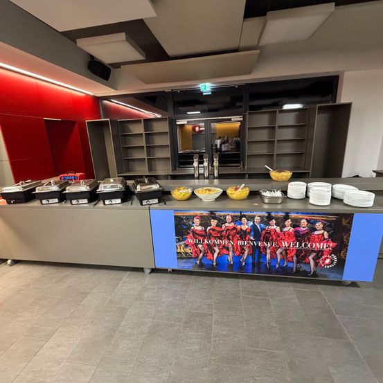 Ein Buffet mit verschiedenen Gerichten, auf einem Banner sind Frauen in roten Kleidern und eine Willkommensbotschaft in mehreren Sprachen abgebildet.