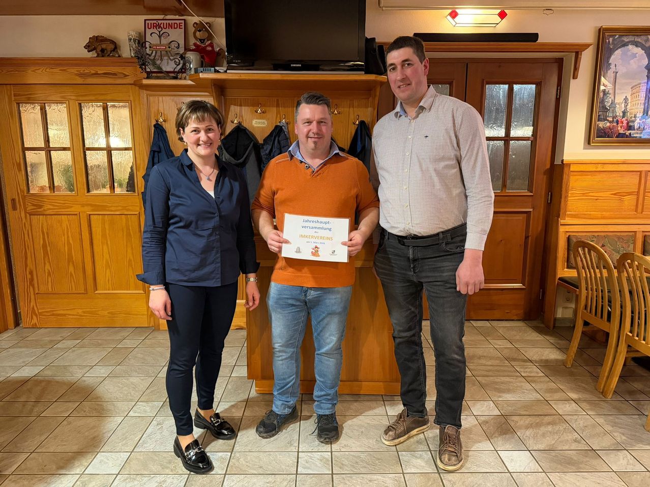 Drei Menschen stehen in einem Raum, zwei Männer und eine Frau. Sie lächeln und posieren für ein Foto. Sie halten ein Zertifikat. Der Mann in der Mitte hält ein Zertifikat. Hinter ihnen steht ein Holzregal.