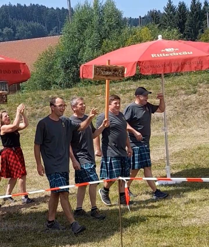 Five people, four men and one woman, are standing in a field with umbrellas and a sign. They are wearing kilts and appear to be posing for a photo.