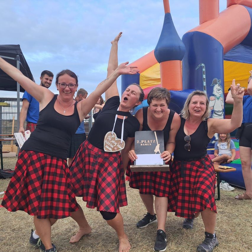 Four women in matching red and black plaid skirts celebrate with a trophy in front of a colorful inflatable castle.