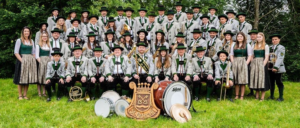 Eine Gruppe von Musikern in traditioneller bayerischer Kleidung posiert für ein Foto auf einer Wiese mit Bäumen im Hintergrund. Sie haben verschiedene Musikinstrumente.