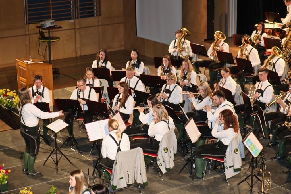 A brass and woodwind band practices on a stage with music stands and sheet music. Members are seated and standing, holding instruments.