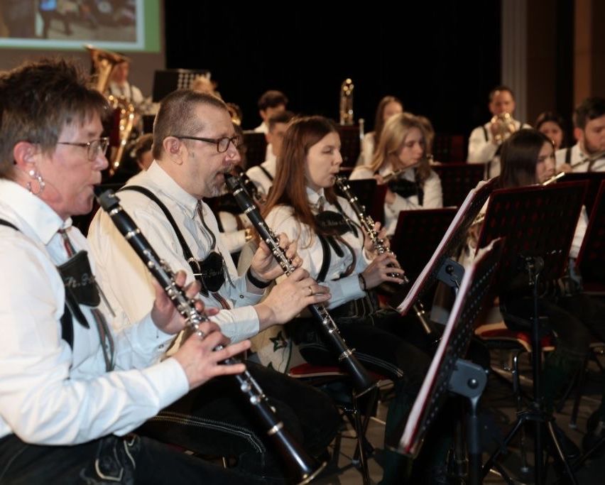 A band practices on stage, with members playing clarinets and trumpets. They wear matching white shirts and suspenders, with a projector screen visible in the background.