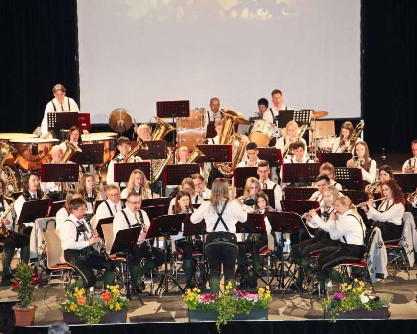 A band performs on a stage with a projector screen in the background. The band members sit in rows with instruments, including trumpets, trombones, and flutes. The stage is adorned with colorful flowers.