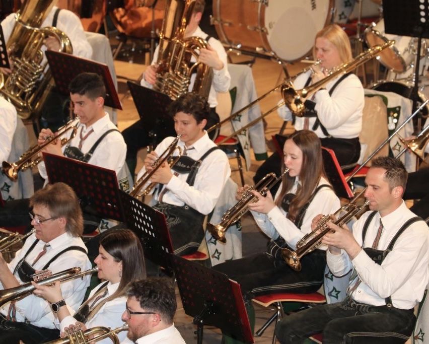 A brass band performs on stage, with members playing trumpets, trombones, and tubas. The musicians wear traditional white shirts and suspenders, and the audience is seated in the background.