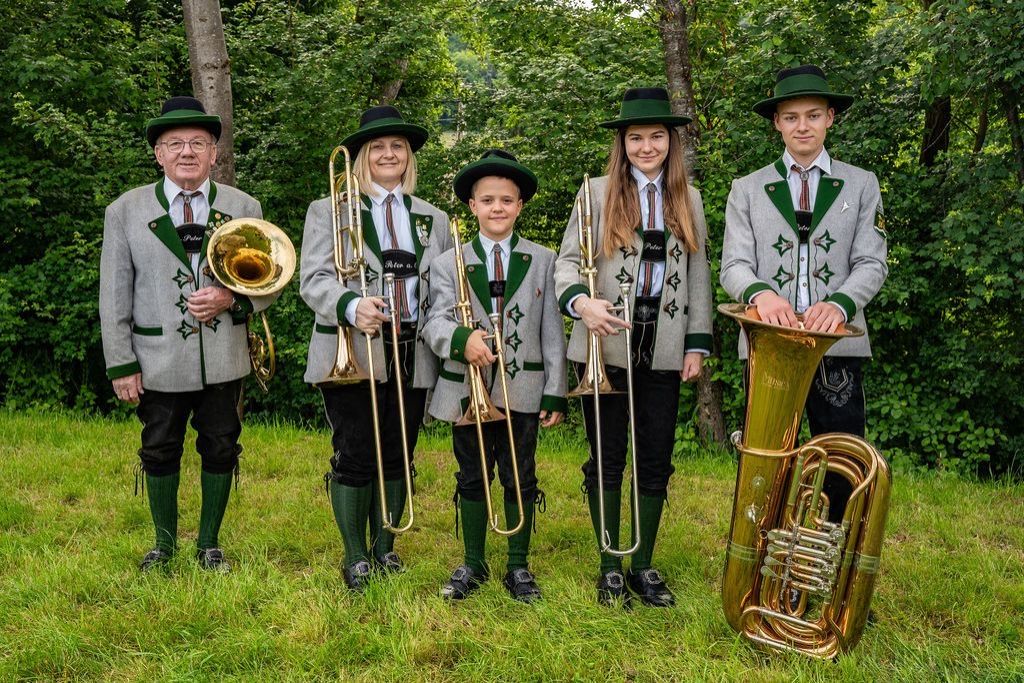 A group of people dressed in traditional attire with musical instruments, posing in a grassy area with trees in the background.