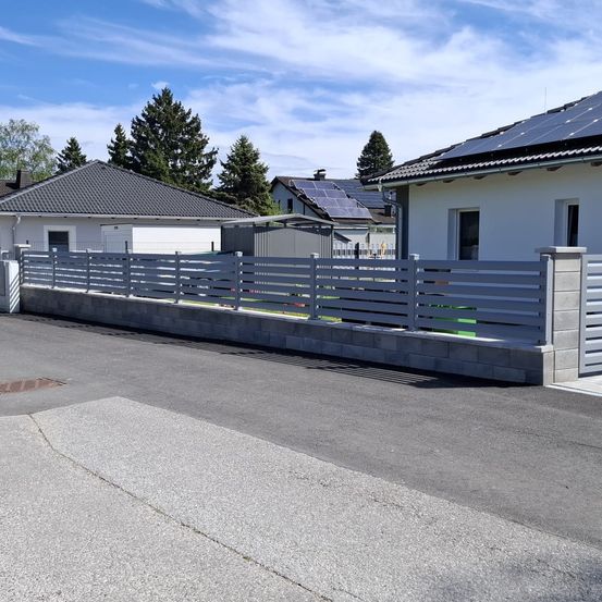 Ein moderner Zaun umgibt eine Einfahrt, mit zwei Häusern und Solarpanelen auf den Dächern im Hintergrund. Bäume und ein klarer Himmel vervollständigen die Szene.
