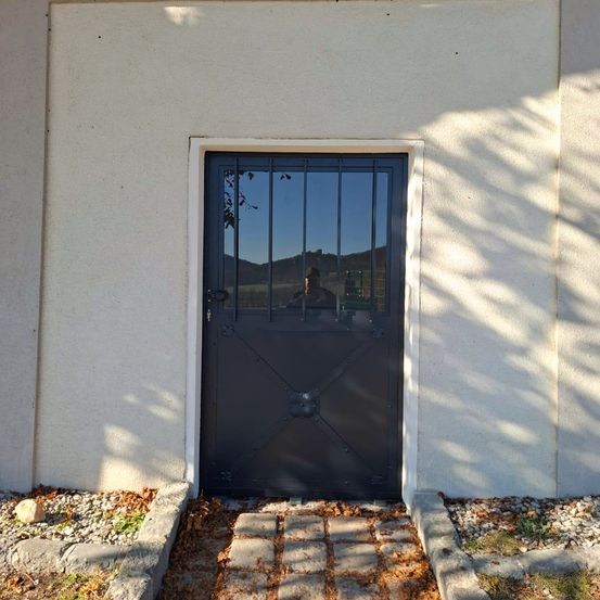 Ein Gebäude mit weißer Wand und schwarzem Eisentor. Das Tor ist geschlossen, und eine Person spiegelt sich im Glasfenster wider.