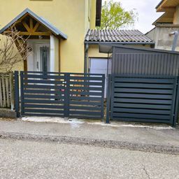 Zwei graue Metallzäune stehen vor einem gelben Gebäude. Das Tor ist offen, mit einem kleinen Stück Gras auf dem Bürgersteig.