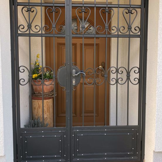 Ein geschlossenes schwarzes Metalltor mit dekorativen Elementen und einer braunen Holztür dahinter. Links neben der Tür stehen zwei Topfpflanzen mit gelben Blumen.