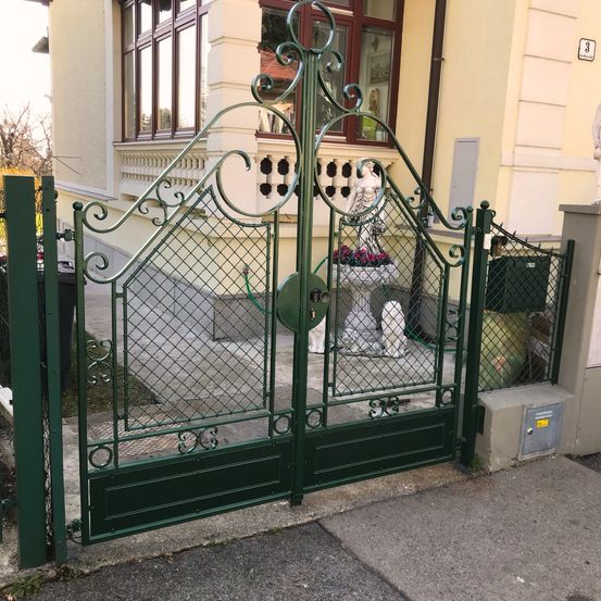 Ein grünes Schmiedeeisentor mit dekorativen Mustern vor einem Gebäude. Eine Statue und Blumen befinden sich hinter dem Tor.