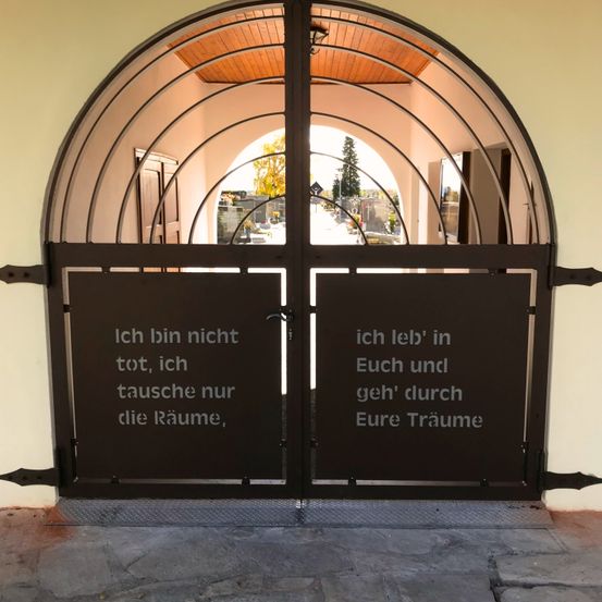 Ein Paar geschwungener schwarzer Tore mit einem Gedicht. Das Gedicht lautet: 'Ich bin nicht tot, ich tausche nur die Räume, ich lebe in Euch und gehe durch Eure Träume.'