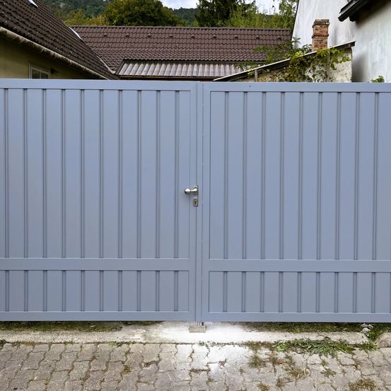 Ein blaues Metalltor mit einem silbernen Griff ist vor einem Haus mit einem braunen Dach und einem Schornstein geschlossen.