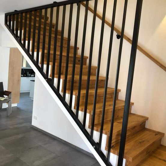 Ein Innenraum einer Wohnung mit einer hölzernen Treppe, die mit einem schwarzen Metallgeländer ausgestattet ist und zu einem Küchenbereich führt.