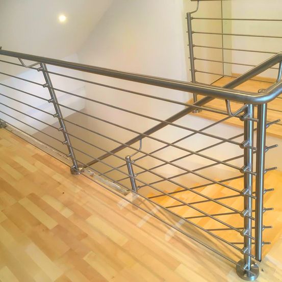 Eine moderne Treppe mit einem Edelstahlgeländer und Holzstufen. Das Geländer weist ein geometrisches Muster auf. Der Raum hat eine weiße Wand und einen Holzboden.