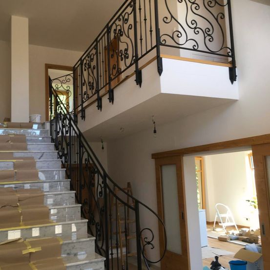 Ein Innenbereich mit einer Marmortreppe, schwarzen Metallgeländern und einer Holztür. Der Raum hat einen Balkon mit einer weißen Wand und einem Holzrahmen.