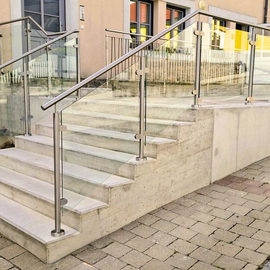 Außenbetonstufen mit Edelstahlgeländer und Glasstufen, die zu einem Gebäude mit Glasfenstern und einem Ziegelpflaster führen.