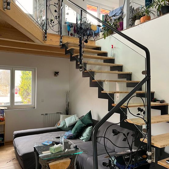 Ein Wohnzimmer mit einem Sofa, einem Glastisch und einer Treppe mit schwarzen Metallgeländern. Ein Fenster mit Blick auf Bäume ist sichtbar.