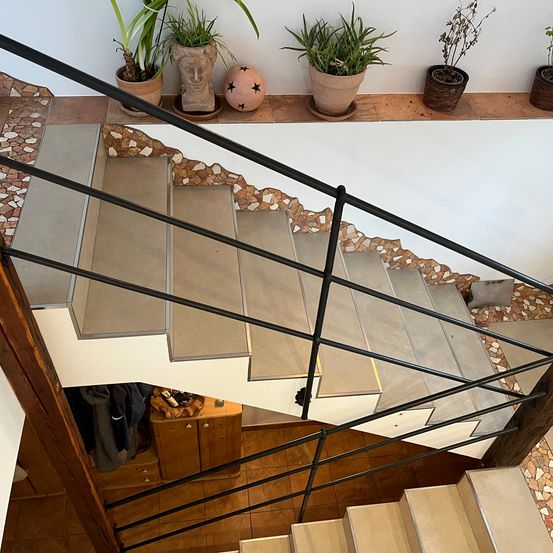 Eine geflieste Treppe mit schwarzem Handlauf, Holzboden und mehreren Topfpflanzen auf einem Regal.