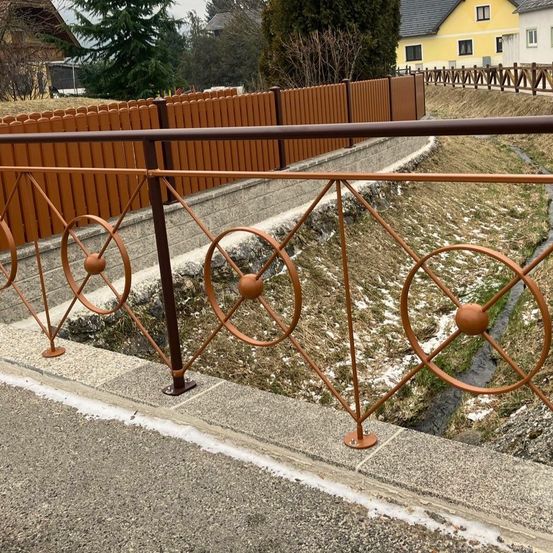 Ein brauner Zaun grenzt eine Straße ab, die zu einem gelben Haus mit Bäumen führt. Der Zaun hat dekorative Elemente.