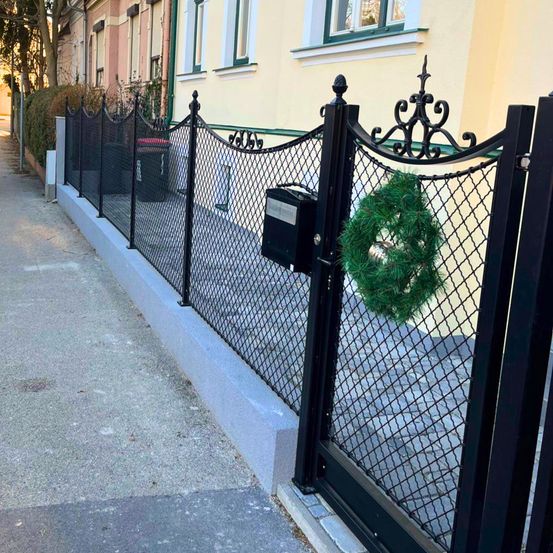 Ein schwarzer Metallzaun grenzt einen Bürgersteig ein. Ein Tor ist offen, mit einem Kranz daran. Ein gelbes Gebäude steht hinter dem Zaun.