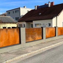 Ein Haus mit einem braunen Dach und Solarpanelen darauf. Es hat eine braune Einfriedung vorn.