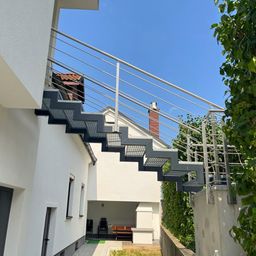 Ein modernes Haus mit einer Metalltreppe und Geländer, mit Blick auf einen Hinterhof mit einer Holzbank und einem Schornstein.