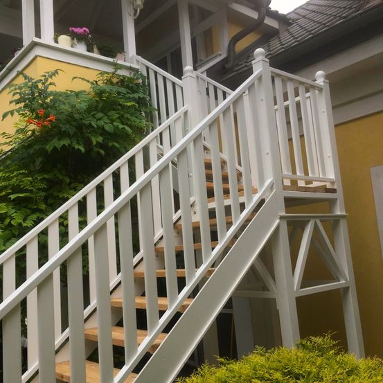 Weiße Holztreppe mit Geländern, die zu einer Veranda eines gelben Gebäudes führt, geschmückt mit grünen Pflanzen und einem Blumentopf.