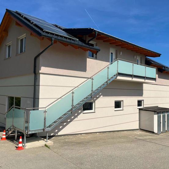 Ein modernes Haus mit Solaranlage auf dem Dach, einer Glastreppe zum Balkon und drei Türen im Erdgeschoss.