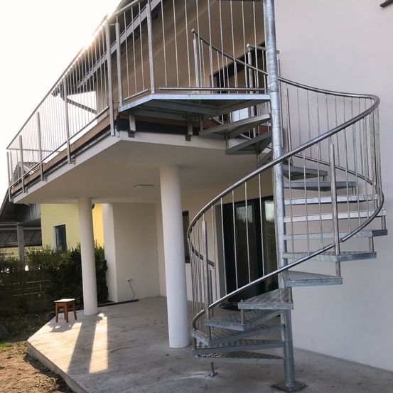 Ein modernes Haus mit einer Wendeltreppe außen, weißen Säulen und einem Balkon mit Metallgeländern. Ein kleiner Holzhocker steht auf dem Patio.