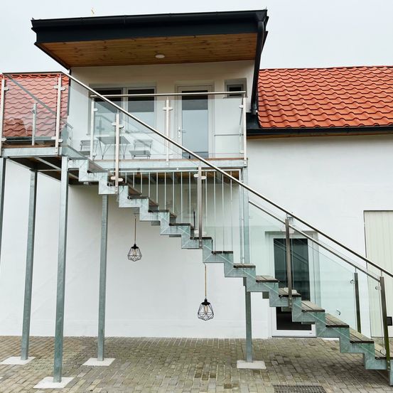 Ein modernes weißes Haus mit Glastreppen und Metallgeländern, die zu einem Balkon mit rotem Dach führen. Der Boden ist aus Ziegelstein und es hängen zwei Lampen.